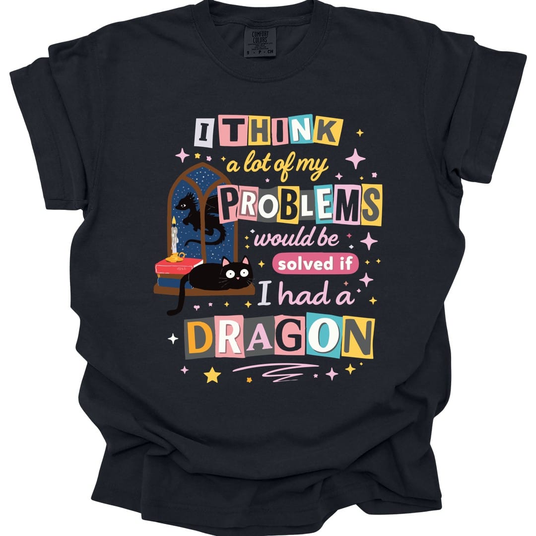 Black t-shirt. Reads "I think a lot of my problems would be solved if I had a dragon" in ransom-note style, each word on its own colored tile in pink, blue, yellow and cream. Behind the words sits a black cat on a windowsill beside a stack of books and an arched window.