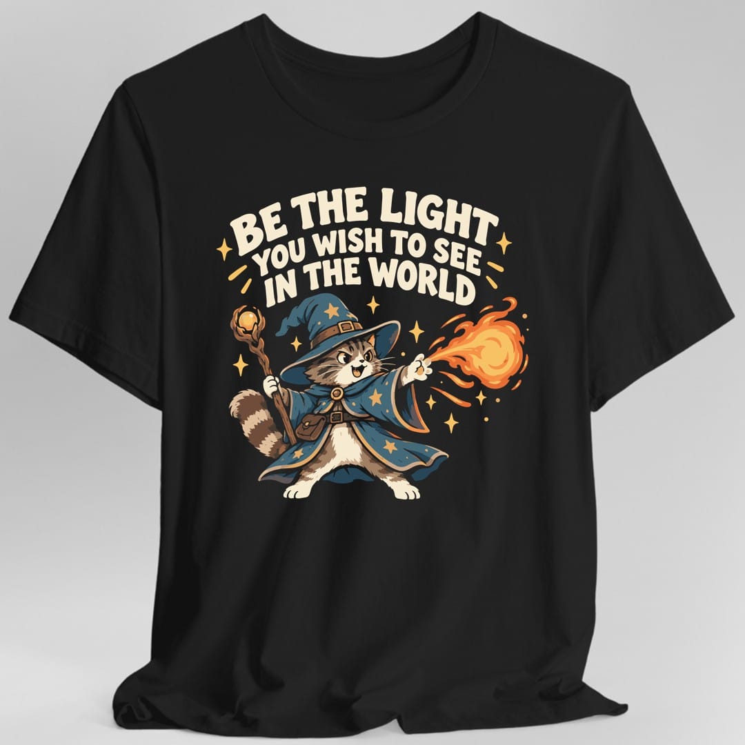 Black t-shirt. The design reads Be The Light You Wish To See In The World in chunky cream capitals arched across the top, with small gold sparkles around the lettering. Below it a brown and white tabby cat stands in a deep blue wizard cloak covered in gold stars and a matching pointed hat, holding a wooden staff topped with a glowing amber orb in one paw and shooting a bright orange fireball from the other. Retro cartoon style.