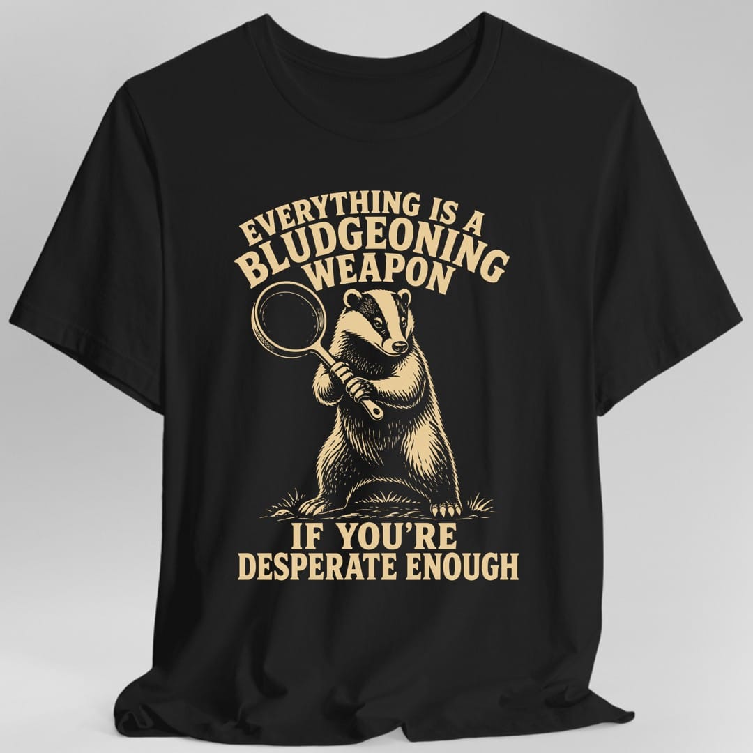 Black t-shirt. The design reads EVERYTHING IS A BLUDGEONING WEAPON in tan capitals arched across the top, and IF YOU'RE DESPERATE ENOUGH in two lines of capitals at the bottom. Between them a badger stands upright in a patch of grass, resting a round cast iron frying pan on its shoulder with both paws. Finely hatched engraving style, all in a single tan ink.