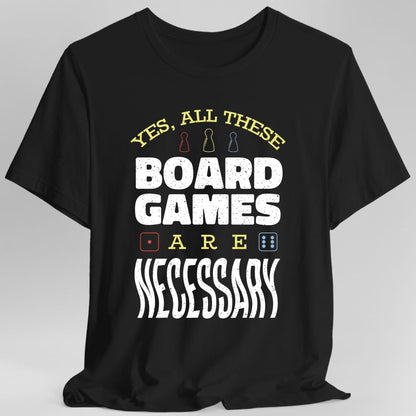 Black t-shirt. Reads "Yes, all these" across the top in gold capitals with three small meeples between the words, then "Board games" in large white capitals, then "Are" with a small red and a small blue die either side, and "Necessary" in large white letters at the bottom.