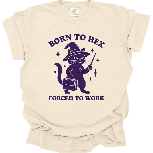 Ivory t-shirt. Reads "Born to Hex" curving across the top and "Forced to Work" across the bottom in purple capitals. Between them a cat in a pointed witch hat stands upright holding a wand in one paw and a briefcase in the other, with small sparkles around it. All in a single deep purple.