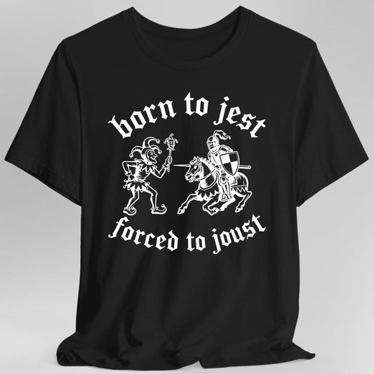 Black t-shirt. The design reads "born to jest" arched across the top and "forced to joust" arched across the bottom, both in blackletter script. Between them a jester in a belled cap and curled shoes leans back waving a marotte, a jester's stick puppet, at an armored knight charging in on a barded horse with a lance and a checkered shield. Woodcut style line art in a single ink.