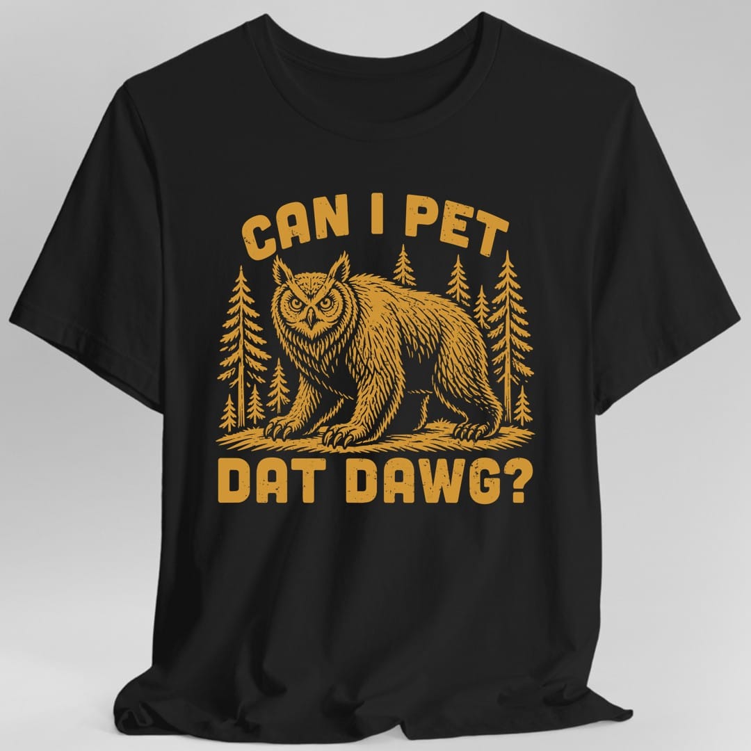 Black t-shirt. The design reads "Can I pet dat dawg?" in chunky arched capitals, split above and below the picture. Between them a shaggy owlbear, part great horned owl and part bear, stands on all fours on a patch of ground with tall pine trees behind it. Retro woodcut style, printed in a single warm gold ink.