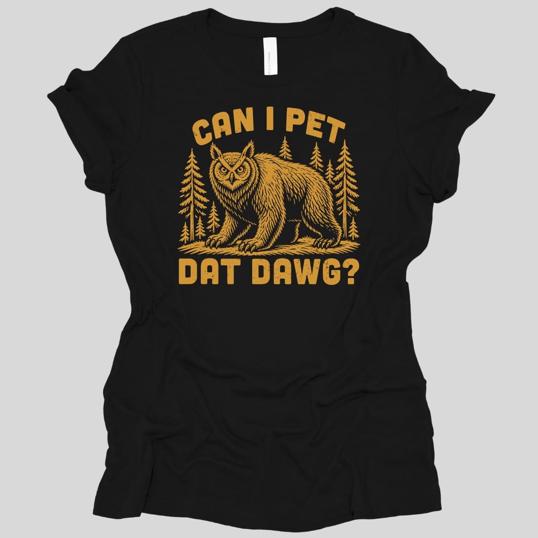 Black women's t-shirt. The design reads "Can I pet dat dawg?" in chunky arched capitals, split above and below the picture. Between them a shaggy owlbear, part great horned owl and part bear, stands on all fours on a patch of ground with tall pine trees behind it. Retro woodcut style, printed in a single warm gold ink.