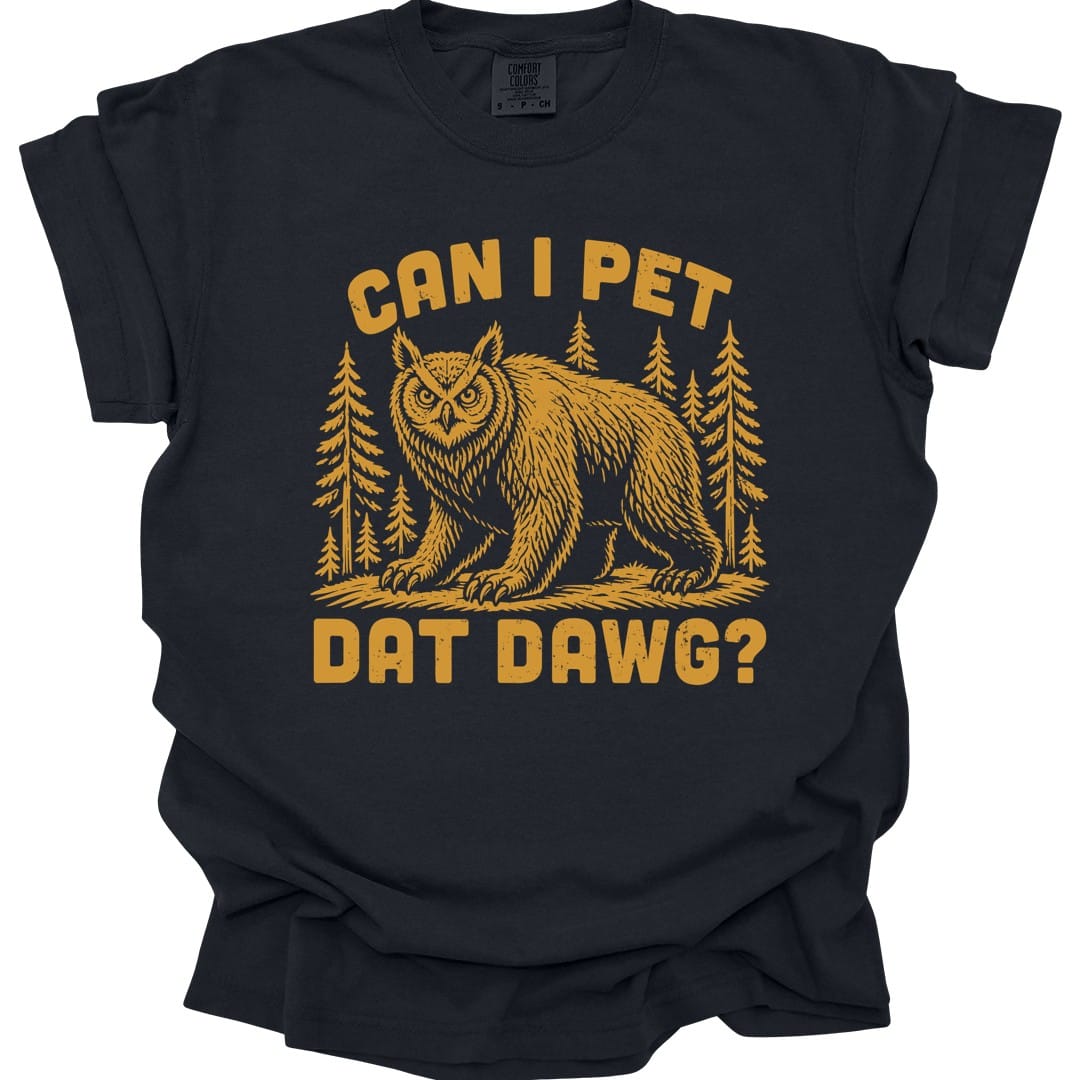Black t-shirt. The design reads "Can I pet dat dawg?" in chunky arched capitals, split above and below the picture. Between them a shaggy owlbear, part great horned owl and part bear, stands on all fours on a patch of ground with tall pine trees behind it. Retro woodcut style, printed in a single warm gold ink.