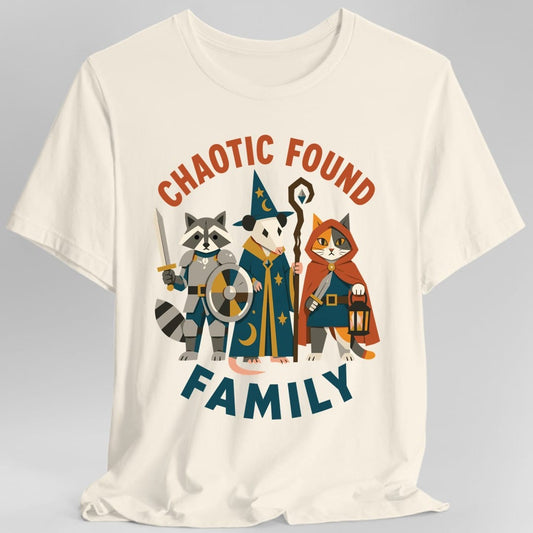 Natural t-shirt. The design reads "CHAOTIC FOUND" in orange letters arched across the top and "FAMILY" in teal across the bottom. Between them three animal adventurers stand shoulder to shoulder in flat illustrated style: a raccoon in grey armor with a sword and round shield, an opossum wizard in a starry teal robe and pointed hat holding a tall staff, and a calico cat in an orange hood with a dagger and a lit lantern.