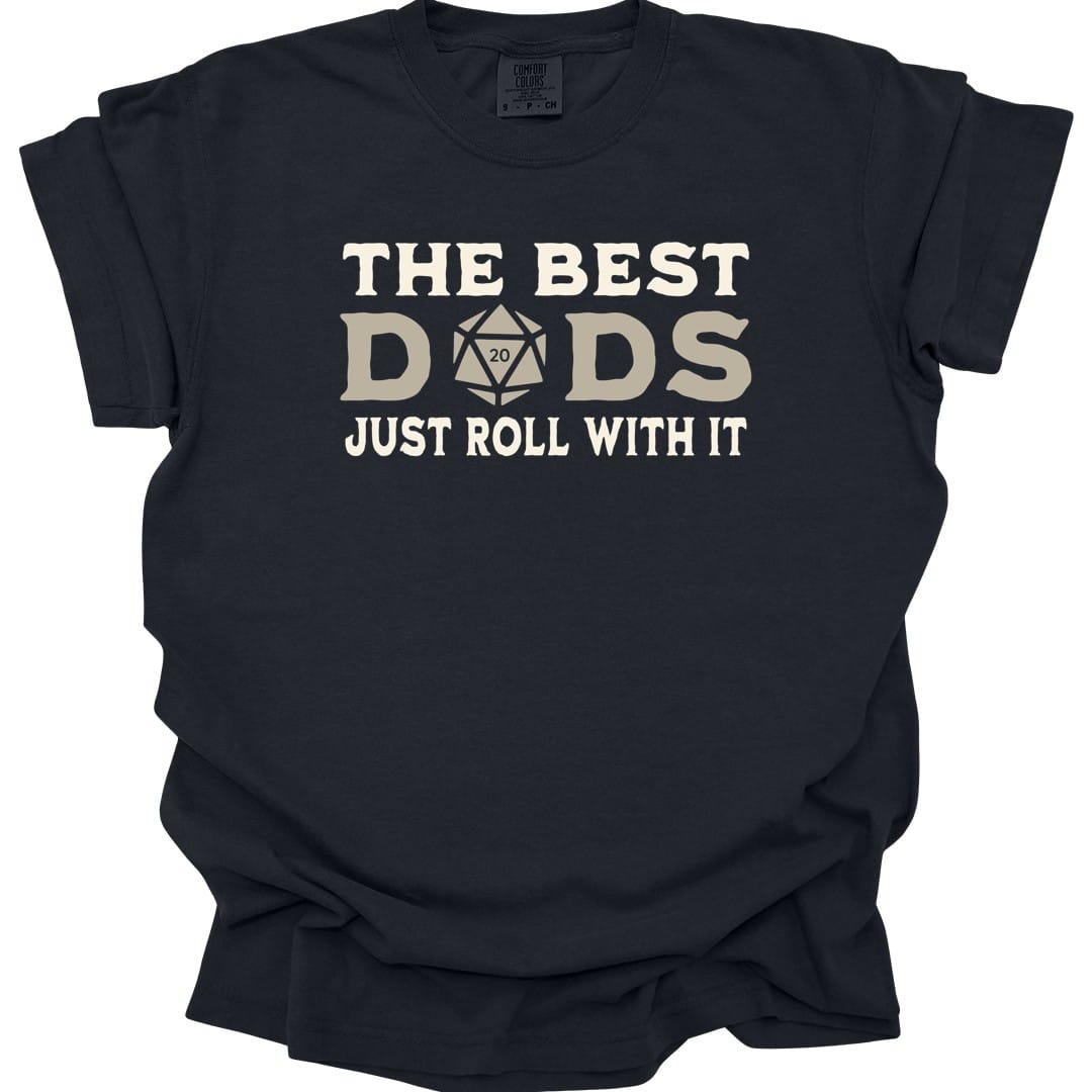 Black t-shirt. Reads "The best" across the top in cream capitals, then "Dads" in large letters where the middle letter is replaced by a twenty-sided die showing 20. Across the bottom, "Just roll with it". All type in cream and tan.