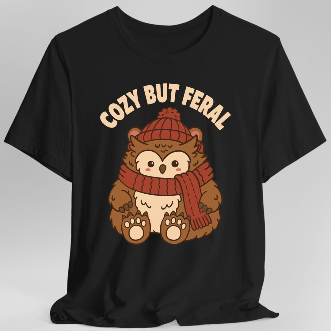 Black t-shirt. The design reads "COZY BUT FERAL" in cream letters arched across the top, above a round cartoon owlbear. It has an owl's face and feathered chest in cream and a brown furry body with big clawed bear paws, and it wears a red knit bobble hat and a long red ribbed scarf wound around its neck. Its wide dark eyes stare straight out. Bold dark brown outlines, soft kawaii illustration in brown, cream and rust red.