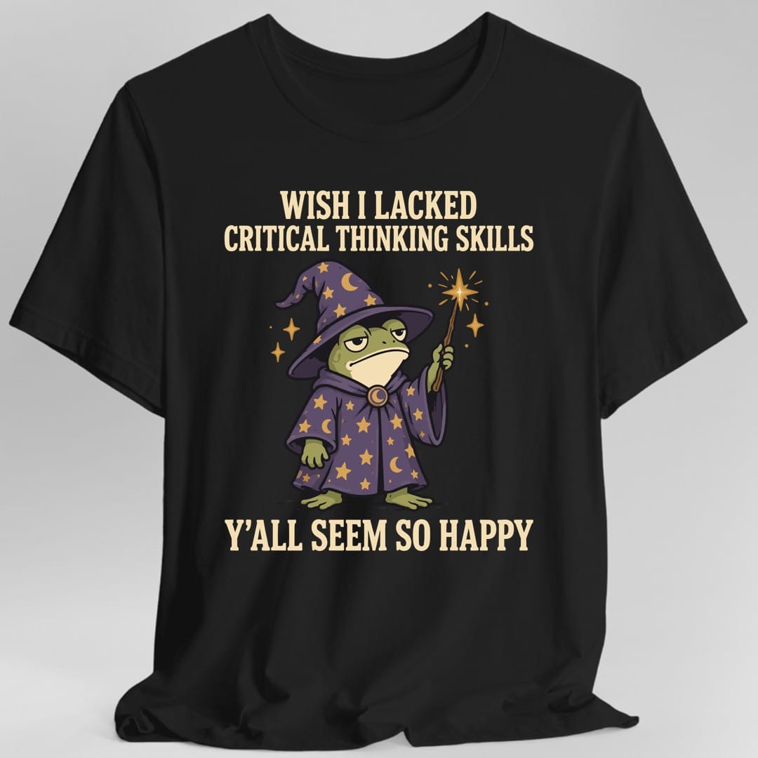 Black t-shirt. Reads "Wish I Lacked Critical Thinking Skills" in bold condensed capitals across the top and "Y'all Seem So Happy" across the bottom. Between them stands a grumpy green frog in a tall purple wizard hat and matching robe covered in gold stars and moons, holding up a wand that throws off a gold spark. Bold outlined cartoon art.