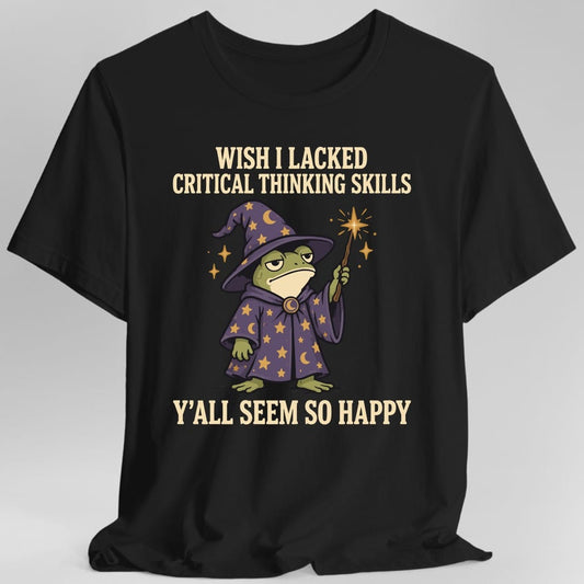 Black t-shirt. Reads "Wish I Lacked Critical Thinking Skills" in bold condensed capitals across the top and "Y'all Seem So Happy" across the bottom. Between them stands a grumpy green frog in a tall purple wizard hat and matching robe covered in gold stars and moons, holding up a wand that throws off a gold spark. Bold outlined cartoon art.