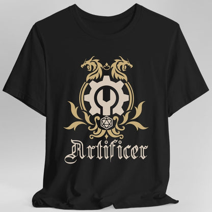 Black t-shirt. A heraldic crest drawn entirely in gold and cream on a plain background. Two dragons face each other over an arched frame, a large cogwheel with a wrench through it sits inside it, and a twenty-sided die hangs below between leafy flourishes. Underneath, "Artificer" in an ornate blackletter script.
