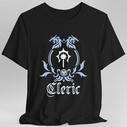 Black t-shirt. A heraldic crest drawn entirely in pale blue and white on a plain background. Two dragons face each other over an arched frame, a spiked mace sits inside the arch, and a twenty-sided die hangs below it between leafy flourishes. Underneath, "Cleric" in an ornate blackletter script.