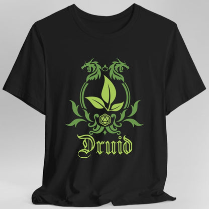 Black t-shirt. A heraldic crest drawn entirely in leaf green on a plain background. Curling vines arch over an arched frame, a sprig of three leaves sits inside it, and a twenty-sided die hangs below between leafy flourishes. Underneath, "Druid" in an ornate blackletter script.
