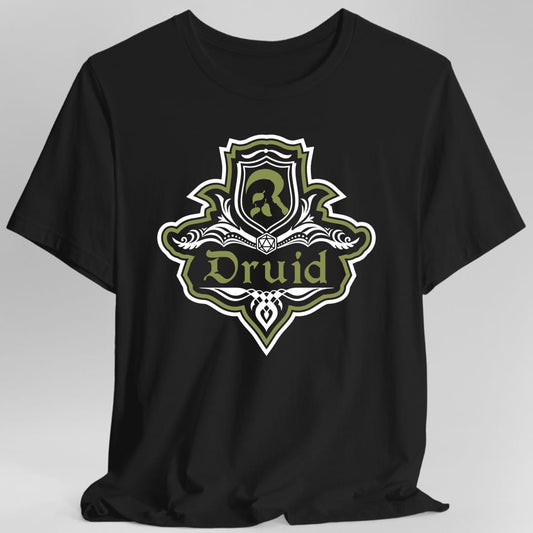 Black t-shirt. A crest shaped like a shield with scalloped edges, outlined in white and olive green. A small inner shield at the top carries an antlered head, an open book with a twenty-sided die on it sits across the middle, and a banner below reads "Druid" in an ornate blackletter script. A pointed pendant hangs from the bottom. White line work with olive green accents.