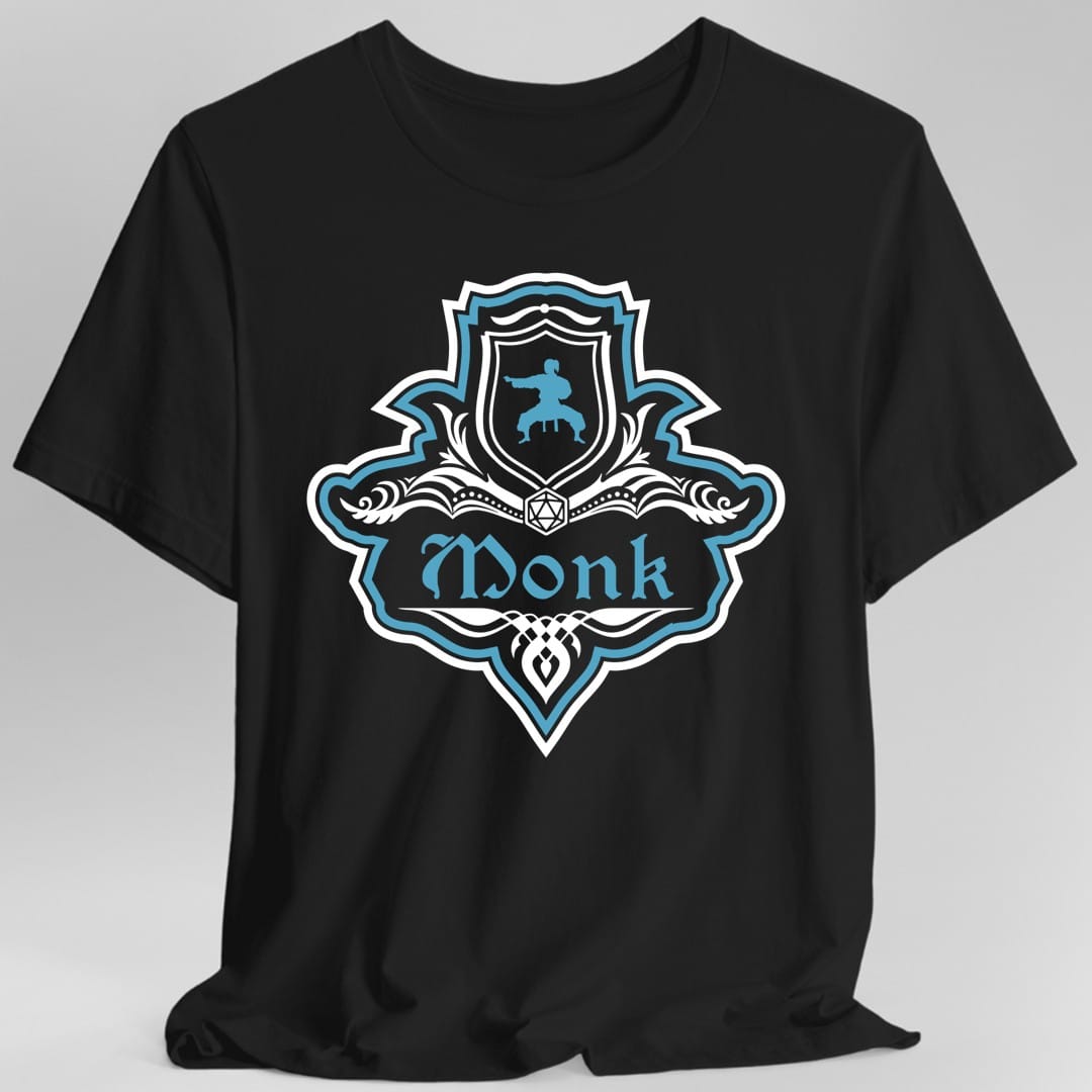 Black t-shirt. A crest shaped like a shield with scalloped edges, outlined in white and bright teal. A small inner shield at the top carries a figure mid martial arts kick, an open book with a twenty-sided die on it sits across the middle, and a banner below reads "Monk" in an ornate blackletter script. A pointed pendant hangs from the bottom. White line work with bright teal accents.