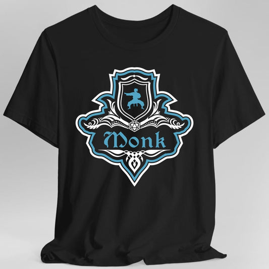 Black t-shirt. A crest shaped like a shield with scalloped edges, outlined in white and bright teal. A small inner shield at the top carries a figure mid martial arts kick, an open book with a twenty-sided die on it sits across the middle, and a banner below reads "Monk" in an ornate blackletter script. A pointed pendant hangs from the bottom. White line work with bright teal accents.