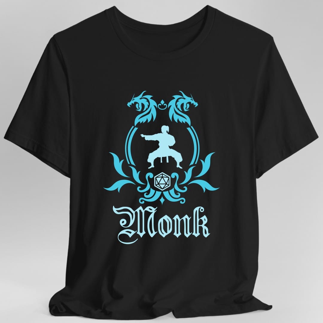 Black t-shirt. A heraldic crest drawn entirely in bright cyan on a plain background. Two dragons face each other over an arched frame, a figure in a martial arts stance sits inside the arch, and a twenty-sided die hangs below it between leafy flourishes. Underneath, "Monk" in an ornate blackletter script.