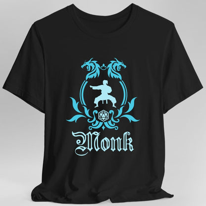 Black t-shirt. A heraldic crest drawn entirely in bright cyan on a plain background. Two dragons face each other over an arched frame, a figure in a martial arts stance sits inside the arch, and a twenty-sided die hangs below it between leafy flourishes. Underneath, "Monk" in an ornate blackletter script.