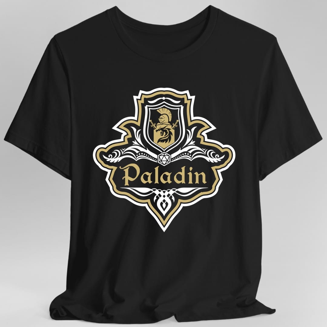 Black t-shirt. A crest shaped like a shield with scalloped edges, outlined in white and gold. A small inner shield at the top carries a plumed helm above a lion, an open book with a twenty-sided die on it sits across the middle, and a banner below reads "Paladin" in an ornate blackletter script. A pointed pendant hangs from the bottom. White line work with gold accents.