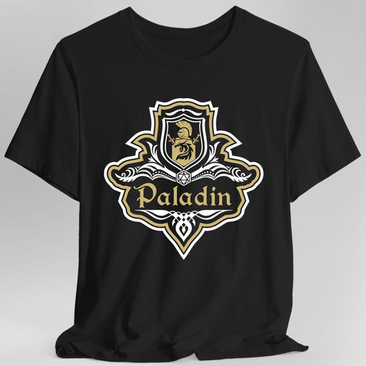 Black t-shirt. A crest shaped like a shield with scalloped edges, outlined in white and gold. A small inner shield at the top carries a plumed helm above a lion, an open book with a twenty-sided die on it sits across the middle, and a banner below reads "Paladin" in an ornate blackletter script. A pointed pendant hangs from the bottom. White line work with gold accents.
