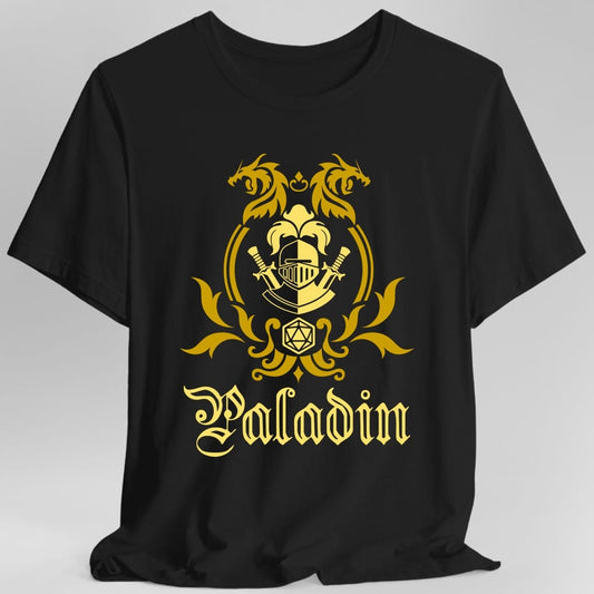 Black t-shirt. A heraldic crest drawn entirely in gold on a plain background. Two dragons face each other over an arched frame, a knight's helm with crossed swords and a fleur de lis sits inside the arch, and a twenty-sided die hangs below it between leafy flourishes. Underneath, "Paladin" in an ornate blackletter script.