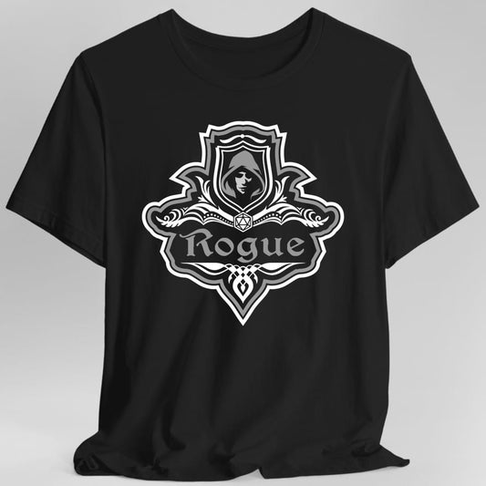 Black t-shirt. A crest shaped like a shield with scalloped edges, outlined in white and gray. A small inner shield at the top carries a hooded face in shadow, an open book with a twenty-sided die on it sits across the middle, and a banner below reads "Rogue" in an ornate blackletter script. A pointed pendant hangs from the bottom. White line work with gray accents.