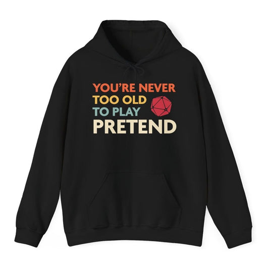 Black hoodie. Reads "You're never too old to play pretend" across four lines, each line in its own color, orange, cream, teal and pale yellow. A red twenty-sided die sits to the right of the third line. All type, no illustration.