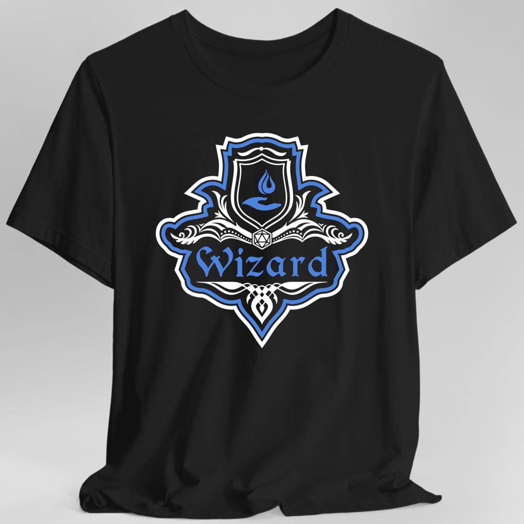 Black t-shirt. A crest shaped like a shield with scalloped edges, outlined in white and royal blue. A small inner shield at the top carries a flame held in a cupped hand, an open book with a twenty-sided die on it sits across the middle, and a banner below reads "Wizard" in an ornate blackletter script. A pointed pendant hangs from the bottom. White line work with royal blue accents.