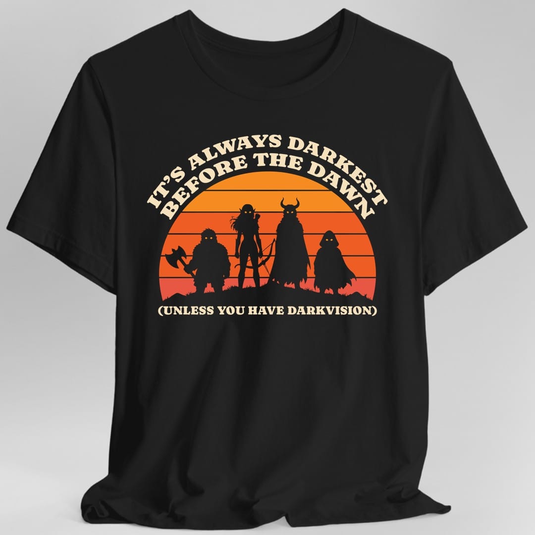 Black t-shirt. The design reads "It's always darkest before the dawn" curved across the top and "(unless you have darkvision)" along the bottom, in cream letters. Between them, four adventurer silhouettes stand shoulder to shoulder on a grassy ridge in front of a big striped sunset that fades from orange at the top down to red. One carries an axe, one a bow, one wears a horned helmet, and one is a hooded figure. Every one of them has glowing gold eyes.