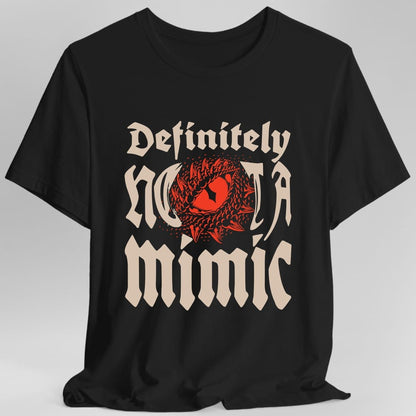 Black t-shirt. Reads "Definitely" in an ornate blackletter script at the top and "Not a mimic" beneath in the same style, all in cream. Behind the middle of the words, a red toothy maw with a single eye opens up, breaking through the lettering.