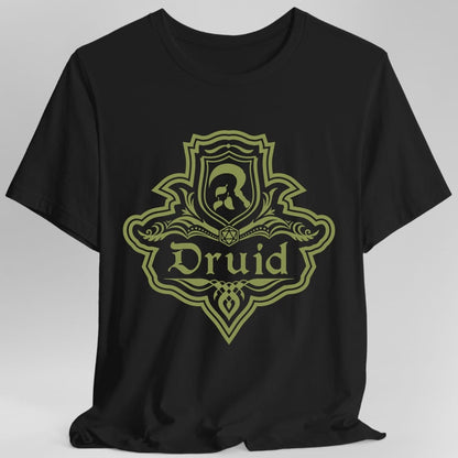 Black t-shirt. A crest shaped like a shield with scalloped edges, drawn entirely in olive green with no other color. A small inner shield at the top carries an antlered head, an open book with a twenty-sided die on it sits across the middle, and a banner below reads "Druid" in an ornate blackletter script. A pointed pendant hangs from the bottom.