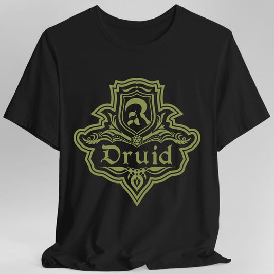 Black t-shirt. A crest shaped like a shield with scalloped edges, drawn entirely in olive green with no other color. A small inner shield at the top carries an antlered head, an open book with a twenty-sided die on it sits across the middle, and a banner below reads "Druid" in an ornate blackletter script. A pointed pendant hangs from the bottom.