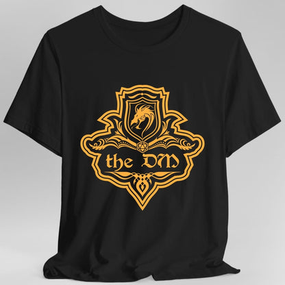 Black t-shirt. A crest shaped like a shield with scalloped edges, drawn entirely in amber orange with no other color. A small inner shield at the top carries a rearing dragon, an open book with a twenty-sided die on it sits across the middle, and a banner below reads "the DM" in an ornate blackletter script. A pointed pendant hangs from the bottom.