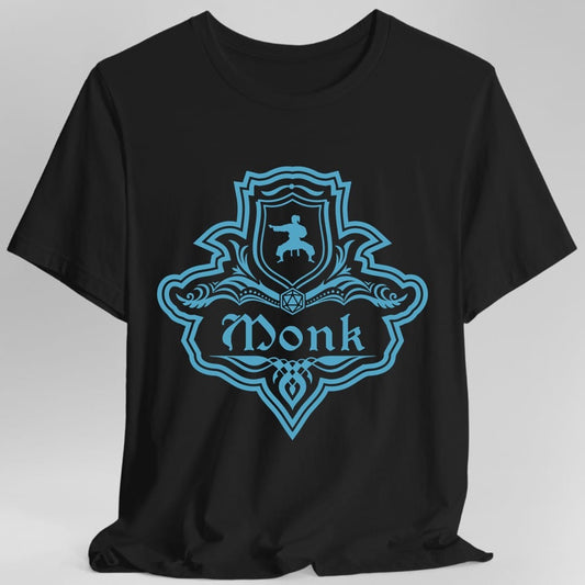 Black t-shirt. A crest shaped like a shield with scalloped edges, drawn entirely in bright teal with no other color. A small inner shield at the top carries a figure mid martial arts kick, an open book with a twenty-sided die on it sits across the middle, and a banner below reads "Monk" in an ornate blackletter script. A pointed pendant hangs from the bottom.