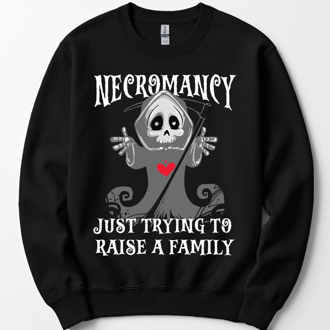 Black sweatshirt. Reads "Necromancy" across the top and "Just trying to raise a family" across the bottom in white spooky capitals. Between them a small cartoon grim reaper in a gray hood holds up both bony hands with a friendly wide-eyed skull face and a red heart on its chest, a scythe leaning behind it. Cute rather than scary. Grays and white with the one red heart.