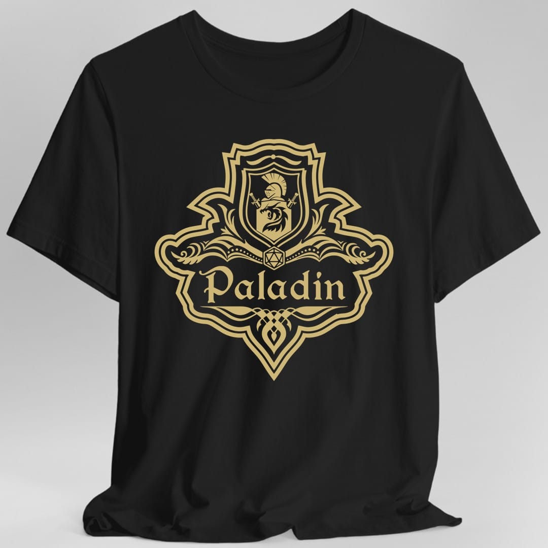 Black t-shirt. A crest shaped like a shield with scalloped edges, drawn entirely in gold with no other color. A small inner shield at the top carries a plumed helm above a lion, an open book with a twenty-sided die on it sits across the middle, and a banner below reads "Paladin" in an ornate blackletter script. A pointed pendant hangs from the bottom.