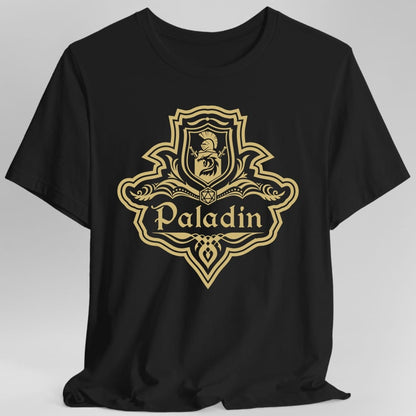 Black t-shirt. A crest shaped like a shield with scalloped edges, drawn entirely in gold with no other color. A small inner shield at the top carries a plumed helm above a lion, an open book with a twenty-sided die on it sits across the middle, and a banner below reads "Paladin" in an ornate blackletter script. A pointed pendant hangs from the bottom.
