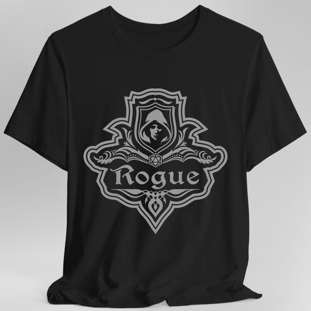 Black t-shirt. A crest shaped like a shield with scalloped edges, drawn entirely in gray with no other color. A small inner shield at the top carries a hooded face in shadow, an open book with a twenty-sided die on it sits across the middle, and a banner below reads "Rogue" in an ornate blackletter script. A pointed pendant hangs from the bottom.
