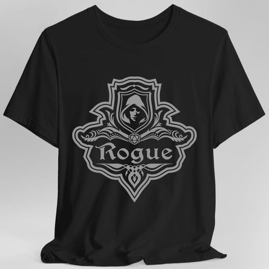 Black t-shirt. A crest shaped like a shield with scalloped edges, drawn entirely in gray with no other color. A small inner shield at the top carries a hooded face in shadow, an open book with a twenty-sided die on it sits across the middle, and a banner below reads "Rogue" in an ornate blackletter script. A pointed pendant hangs from the bottom.