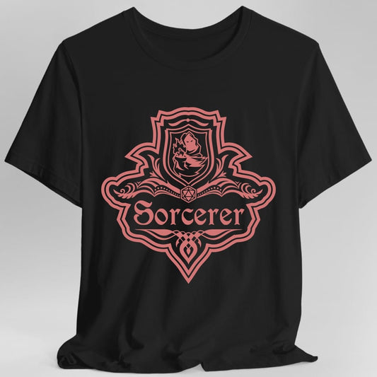 Black t-shirt. A crest shaped like a shield with scalloped edges, drawn entirely in coral red with no other color. A small inner shield at the top carries a dragon curled around a flame, an open book with a twenty-sided die on it sits across the middle, and a banner below reads "Sorcerer" in an ornate blackletter script. A pointed pendant hangs from the bottom.