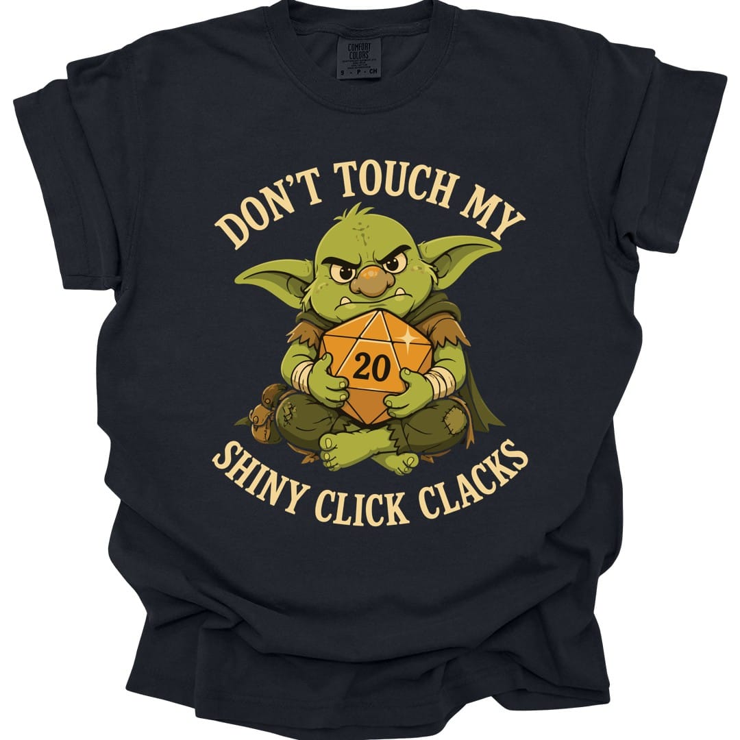 Black t-shirt. Reads "Don't touch my" curving across the top and "Shiny click clacks" across the bottom in cream capitals. Between them a green goblin sits scowling and hugging a large orange twenty-sided die showing 20 to its chest with both arms.