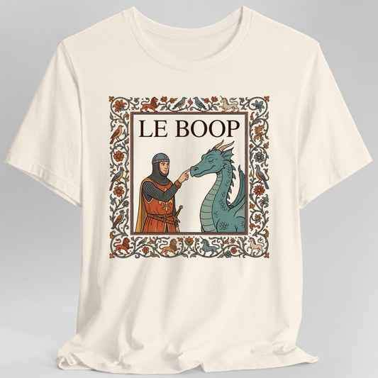 Natural t-shirt. The design reads "Le Boop" in serif capitals at the top of a framed panel, styled like a page from an illuminated manuscript. Inside the panel a knight in chainmail and an orange fleur-de-lis surcoat reaches out and touches the snout of a green dragon, which has its eyes closed. A thick decorated border of vines, orange and blue flowers, lions, unicorns and birds runs all the way around. Medieval manuscript art in orange, teal, gold and brown.
