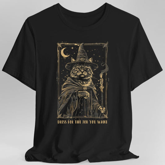 Black t-shirt. The design reads "DRESS FOR THE JOB YOU WANT" in gold capitals along the bottom. Above it, drawn as a gold tarot card with a decorative border, a long-haired cat sits facing forward in a tall pointed wizard hat and a flowing cloak. A crescent moon and scattered stars fill the upper left and a lit candle stands at the right. Fine gold and soft grey line work throughout.