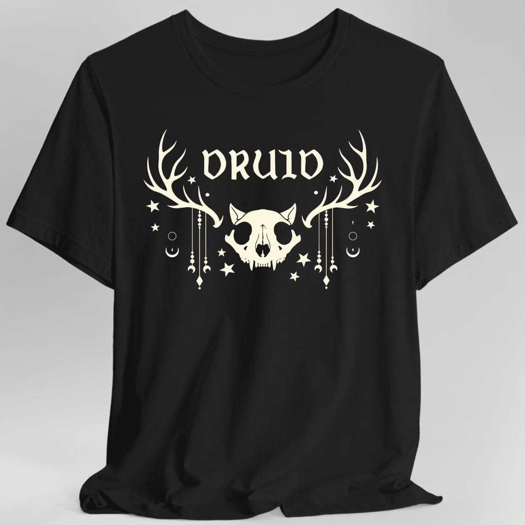 Black t-shirt. The design reads "DRUID" in tall pointed gothic letters across the top. Below it sits an animal skull with pointed ears and fangs, with a huge set of antlers spreading out on either side. Strings of beads and crescent moons hang down from the antlers, and small stars, dots and circles are scattered around the whole thing. All of it is drawn in a single soft cream color, flat and symmetrical.