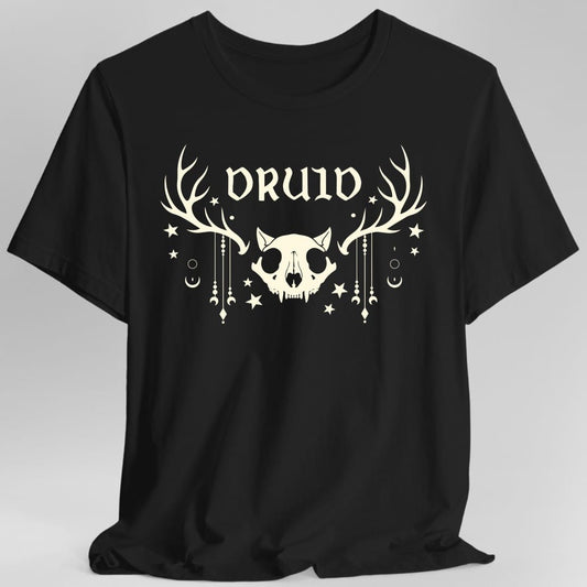 Black t-shirt. The design reads "DRUID" in tall pointed gothic letters across the top. Below it sits an animal skull with pointed ears and fangs, with a huge set of antlers spreading out on either side. Strings of beads and crescent moons hang down from the antlers, and small stars, dots and circles are scattered around the whole thing. All of it is drawn in a single soft cream color, flat and symmetrical.
