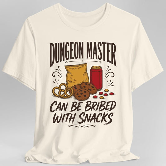 Natural t-shirt. The design reads DUNGEON MASTER across the top and CAN BE BRIBED WITH SNACKS across the bottom in tall vintage lettering with curled flourishes. Between them sits a spread of snacks, a bag of chips, a red soda can, chocolate chip cookies, a soft pretzel and scattered candies. Drawn in warm retro label style.