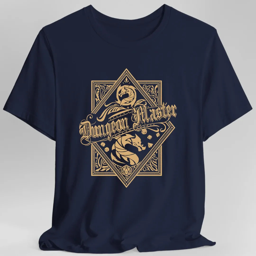 Navy t-shirt. An ornate gold crest, all fine line work on a plain background. A diamond sits over a square frame, both edged with intricate patterned borders. A dragon curls in a roundel at the top, a ribbon banner sweeps across the middle reading "Dungeon Master" in ornate blackletter, and a coiled dragon sits below it among scattered polyhedral dice. A twenty-sided die marks the bottom point.