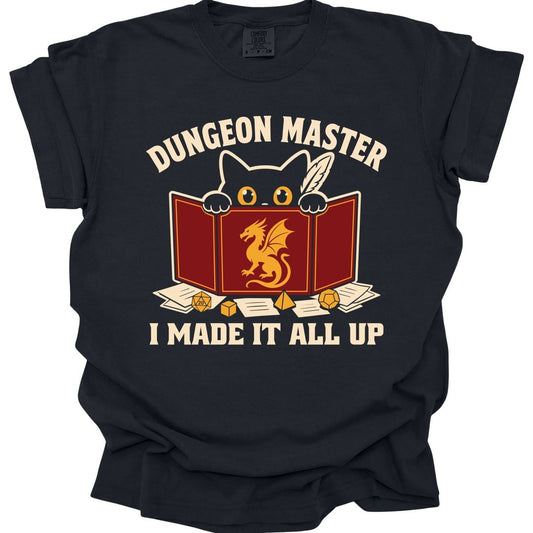 Black t-shirt. Reads "Dungeon Master" curving across the top in cream capitals. Below, a white cat peers over the top of a dark red dungeon master screen with a gold dragon printed on it, paws hooked over the edge, papers and dice scattered around. Across the bottom, "I made it all up".