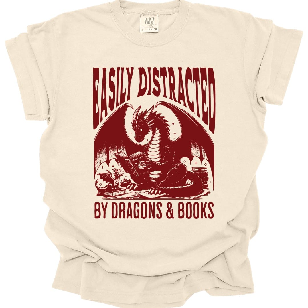 Ivory t-shirt. Reads "Easily distracted" across the top and "By dragons and books" across the bottom in tall dark red capitals. Between them a dragon lies curled up reading an open book, surrounded by more stacked books and scrolls. Engraved line work, all in one deep red.