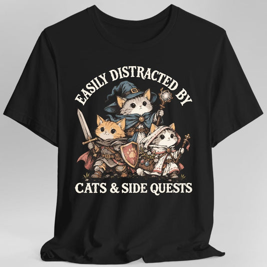 Black t-shirt. Reads "Easily Distracted By" in an arch across the top and "Cats & Side Quests" across the bottom. Between them stand three kitten adventurers: a gray and white wizard in a blue pointed hat and cloak holding a glowing crystal staff, an orange tabby fighter in leather armor with a raised sword and a shield painted with a paw print, and a white robed cleric with a flower satchel and a staff. Soft painted illustration.